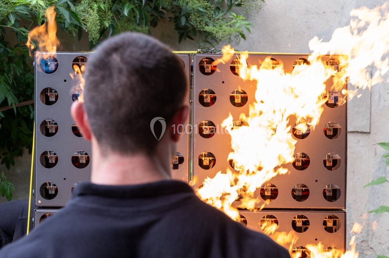 Un homme de dos observe un mur de dispositifs avec des flammes jaillissant à l'extérieur, entouré de végétation.