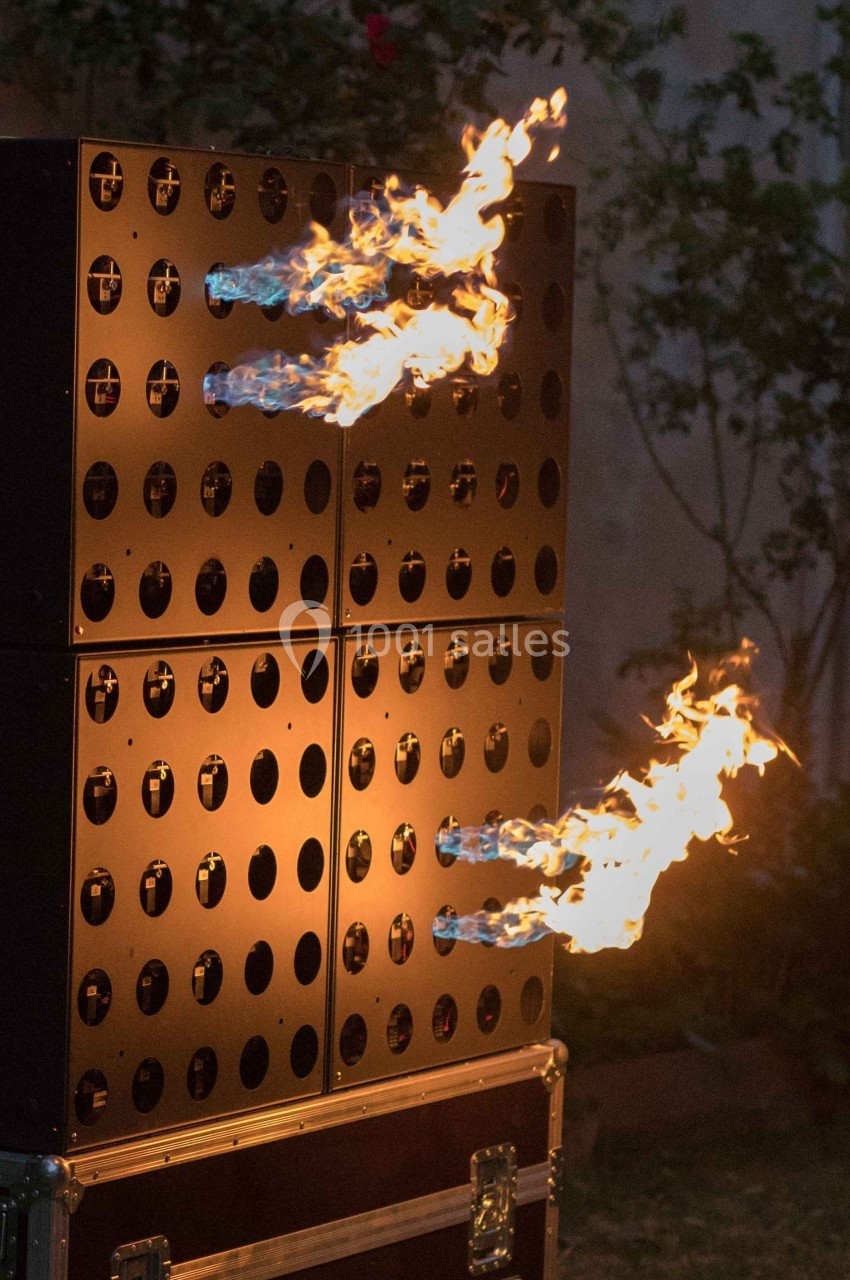Mur de haut-parleurs avec des flammes jaillissant de certains orifices, en extérieur, dans un décor végétal.