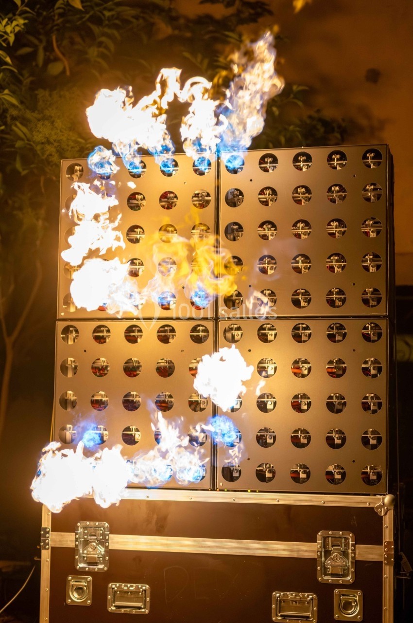 Mur de panneaux métalliques avec des flammes jaillissant, éclairé dans un environnement extérieur nocturne.