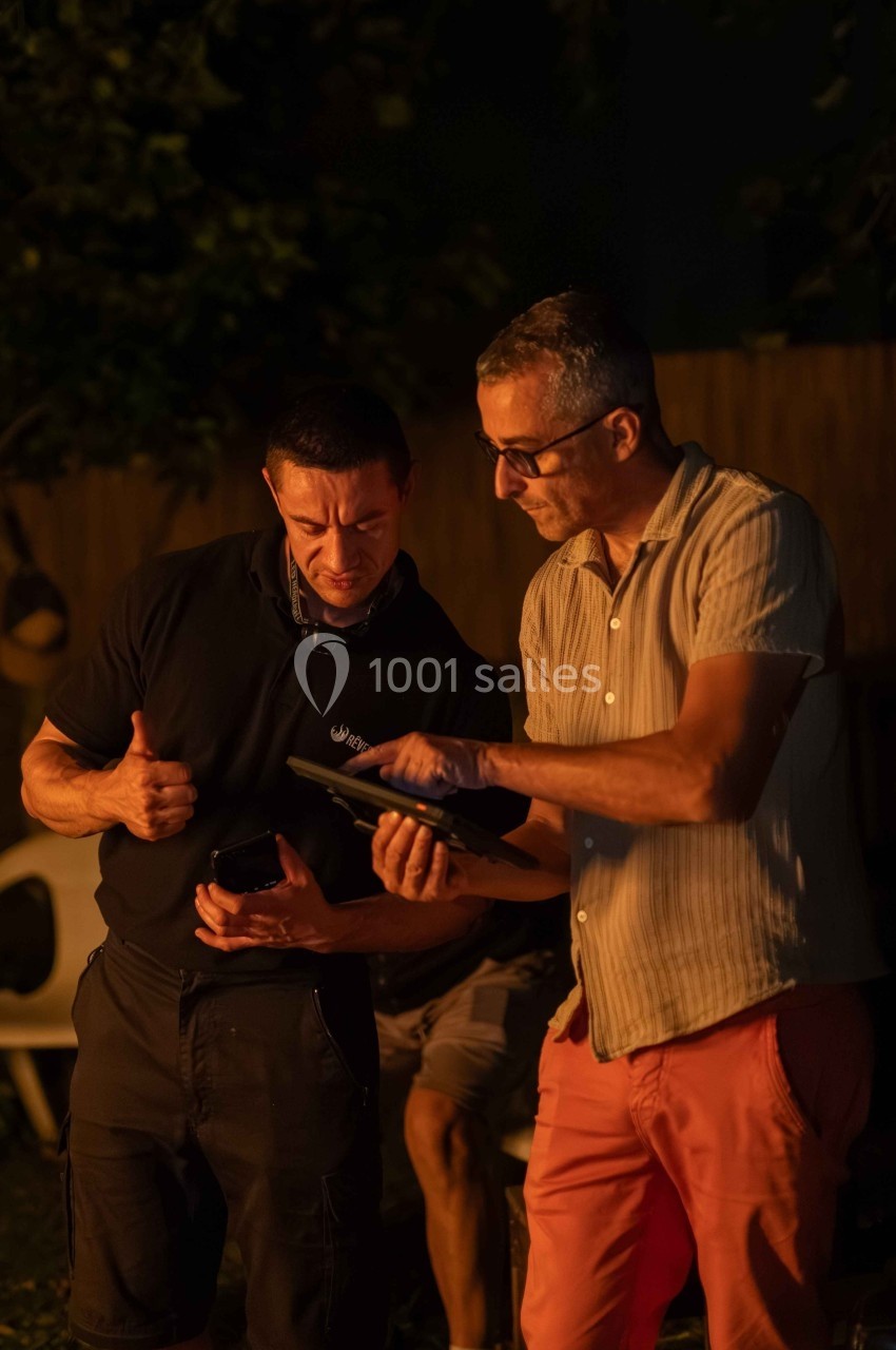 Deux hommes discutent et regardent une tablette dans un jardin, éclairés par une lumière chaude en soirée.