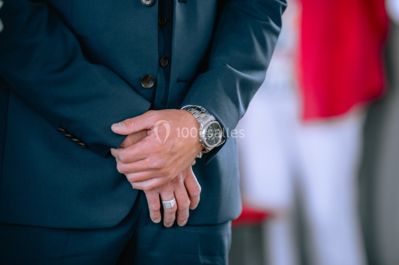 Homme en costume bleu marine, mains croisées, portant une montre argentée et une bague argentée.