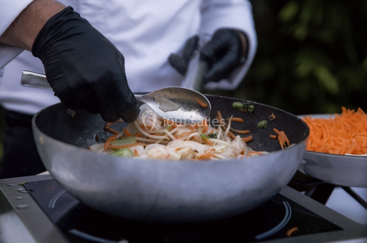 Un cuisinier mélange des légumes sautés dans un wok avec une cuillère en métal, portant des gants noirs.