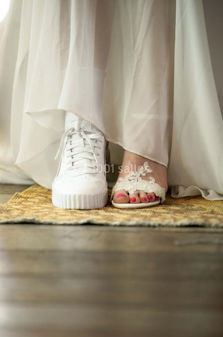 Une mariée porte une basket blanche et une sandale ornée, posées sur un tapis en fibre naturelle.