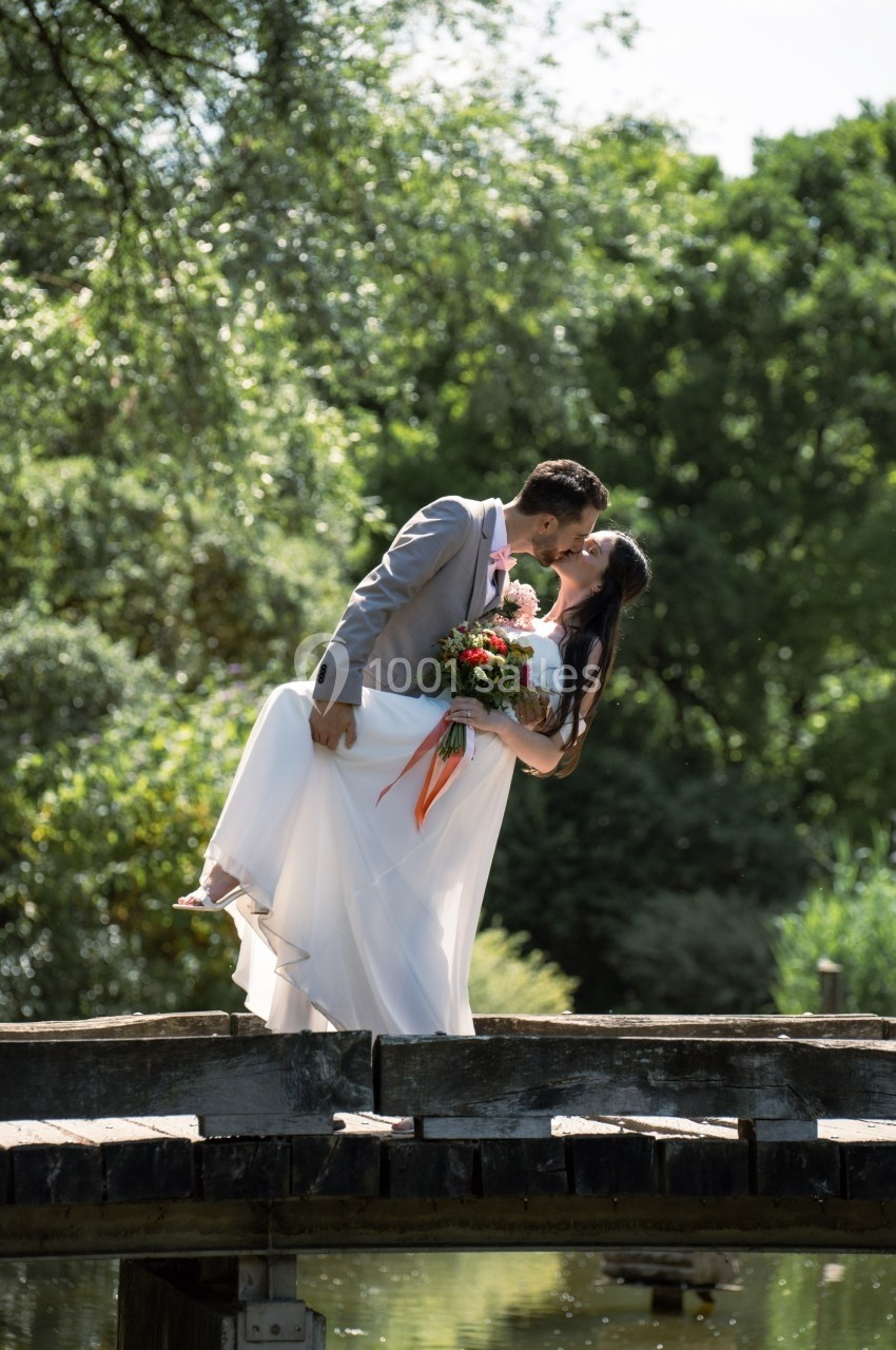 Un couple en tenue de mariage s'embrasse sur un pont en bois, entouré de verdure et baigné de lumière naturelle.