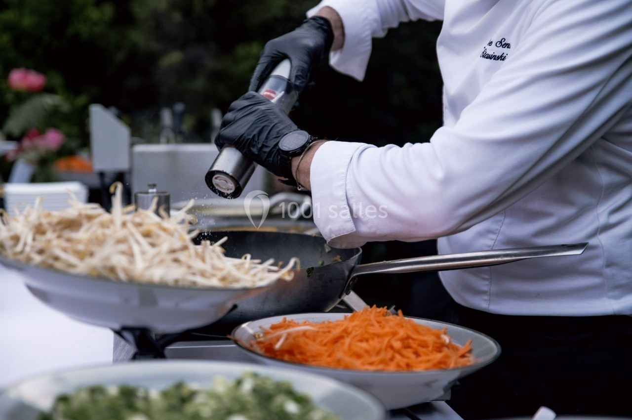 Un chef en tenue blanche assaisonne un plat dans une poêle, entouré de bols de légumes frais.