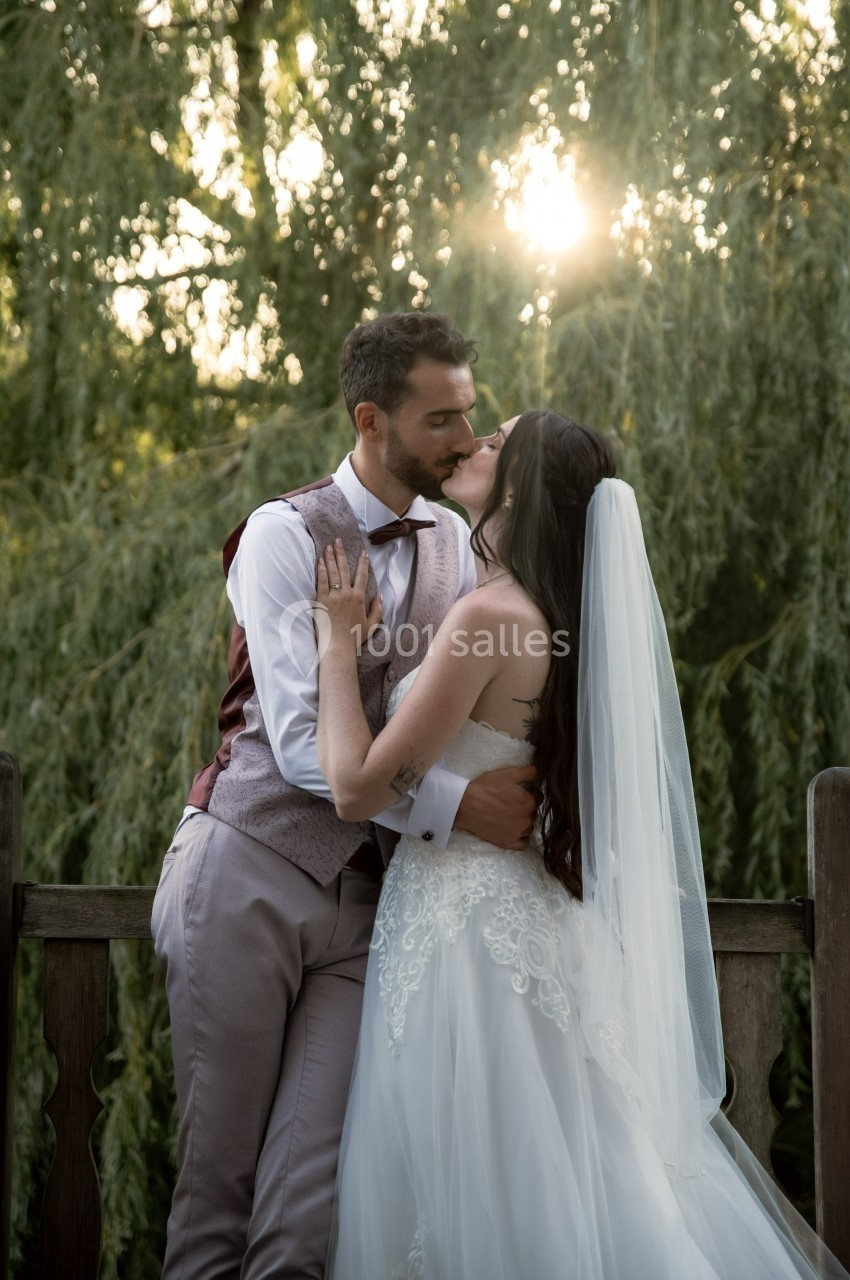 Un couple en tenue de mariage s'embrasse tendrement sur un pont en bois, entouré de verdure et de lumière naturelle.