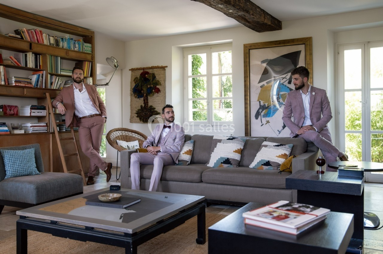Trois hommes en costumes pastel posent dans un salon lumineux avec canapé, table basse et bibliothèque en arrière-plan.