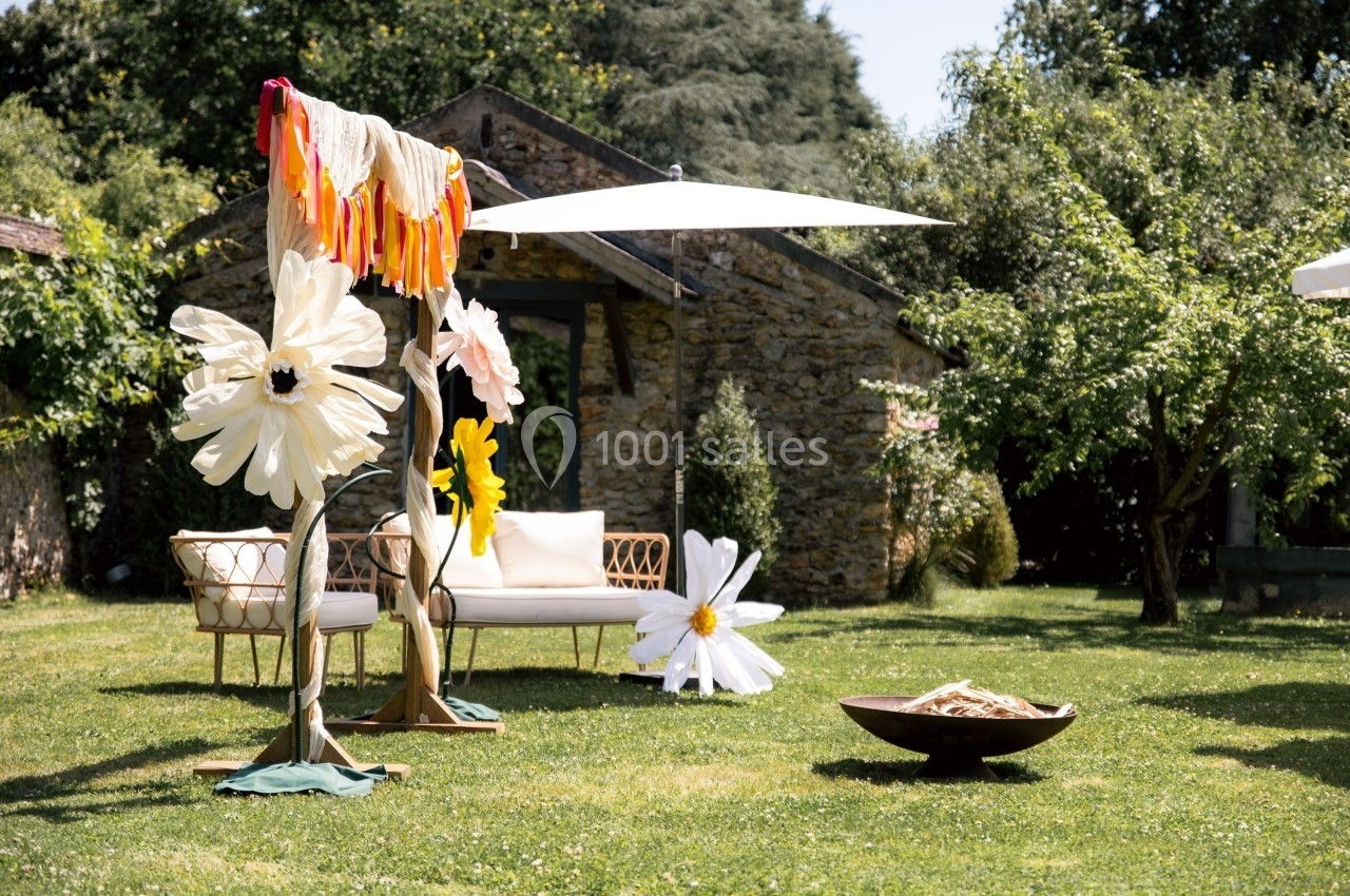 Décoration extérieure avec grandes fleurs en papier, mobilier en rotin et parasol, dans un jardin verdoyant.