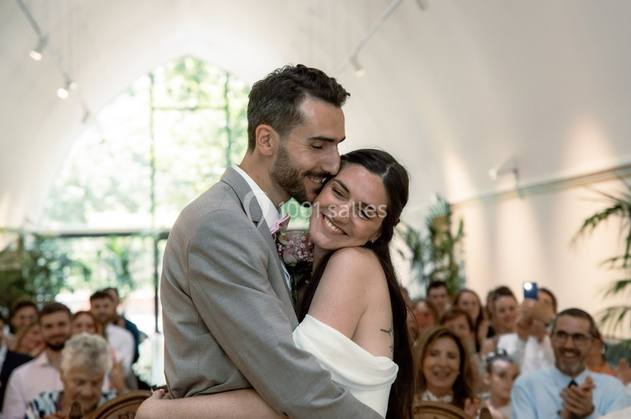Un couple souriant s'enlace lors d'une cérémonie de mariage dans une salle lumineuse avec des invités en arrière-plan.