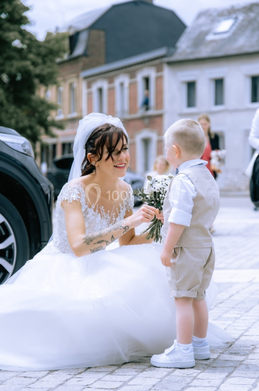 Une mariée accroupie sourit à un jeune garçon en tenue claire, tenant un bouquet de fleurs, dans une rue pavée.