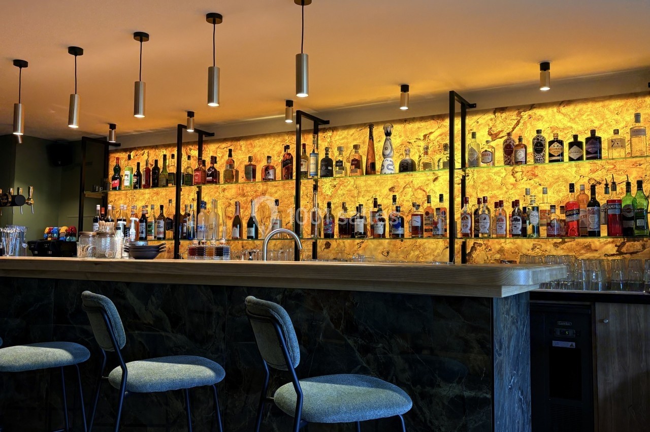 Bar moderne avec comptoir en pierre sombre, tabourets rembourrés et étagères illuminées présentant des bouteilles d'alcool.