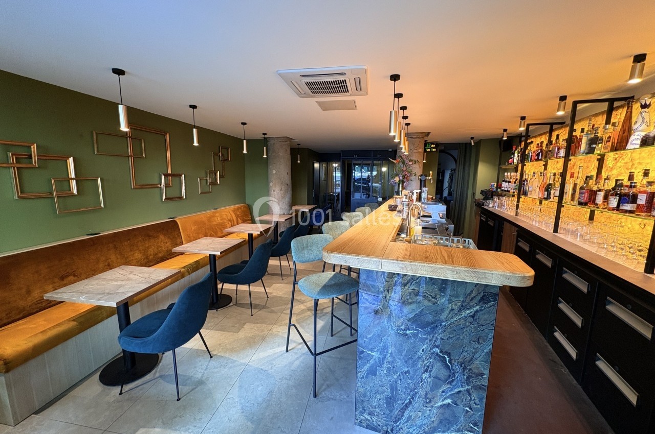 Intérieur d'un bar moderne avec comptoir en marbre, banquettes, chaises et étagères de bouteilles illuminées.