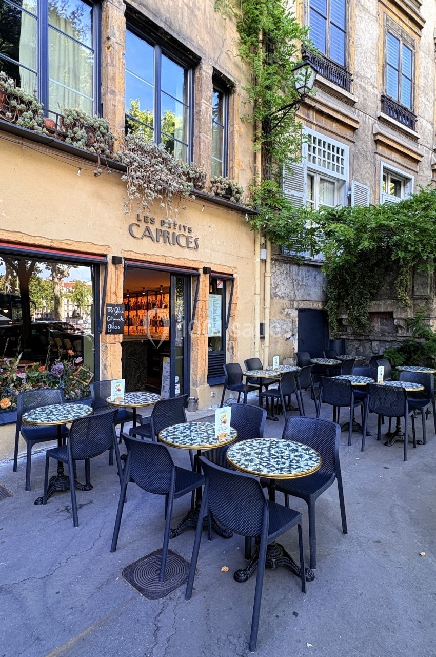 Terrasse d'un café avec des tables rondes et chaises noires, située devant une façade en pierre ornée de plantes.
