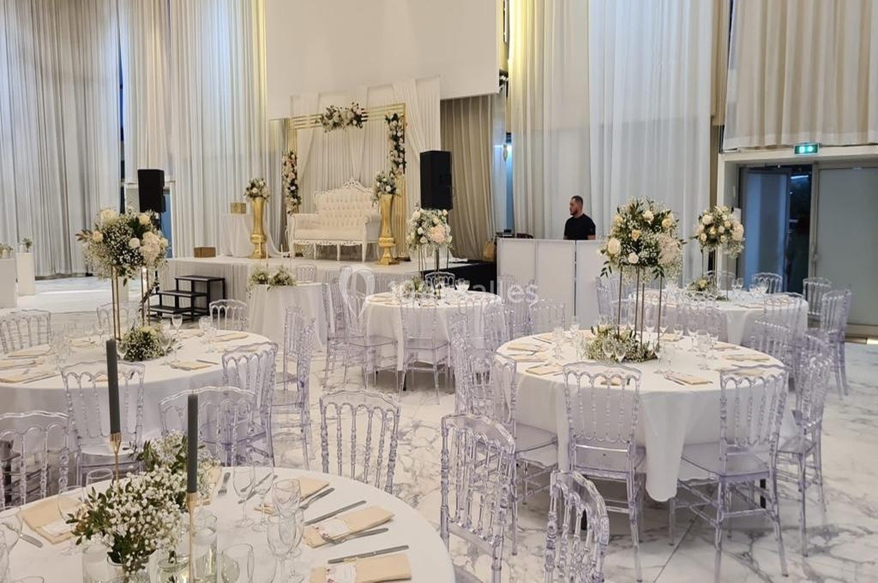 Salle de réception élégante avec tables rondes dressées, chaises transparentes et décorations florales blanches.