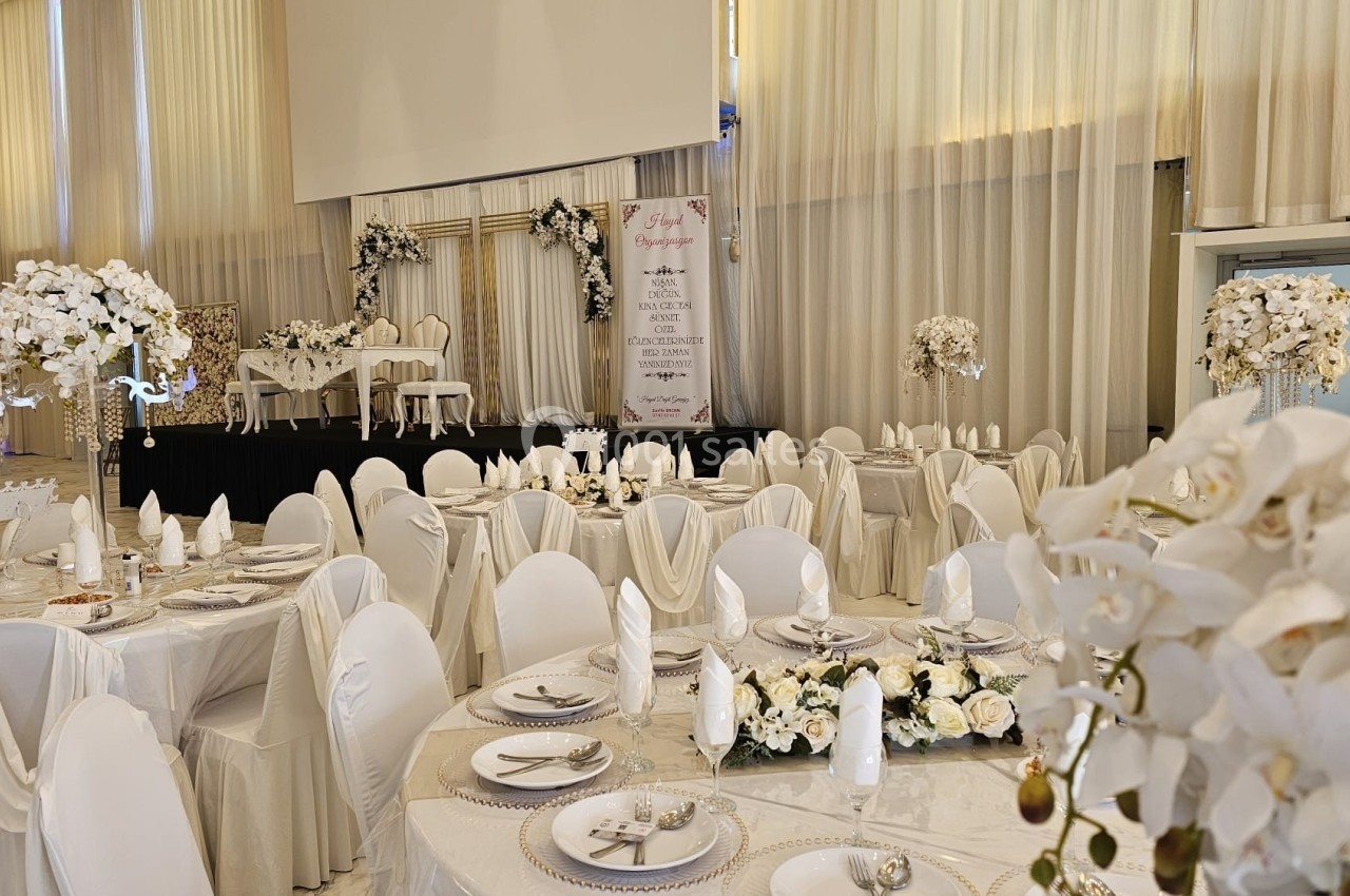 Salle de réception décorée en blanc avec tables dressées, chaises couvertes et arrangements floraux élégants.
