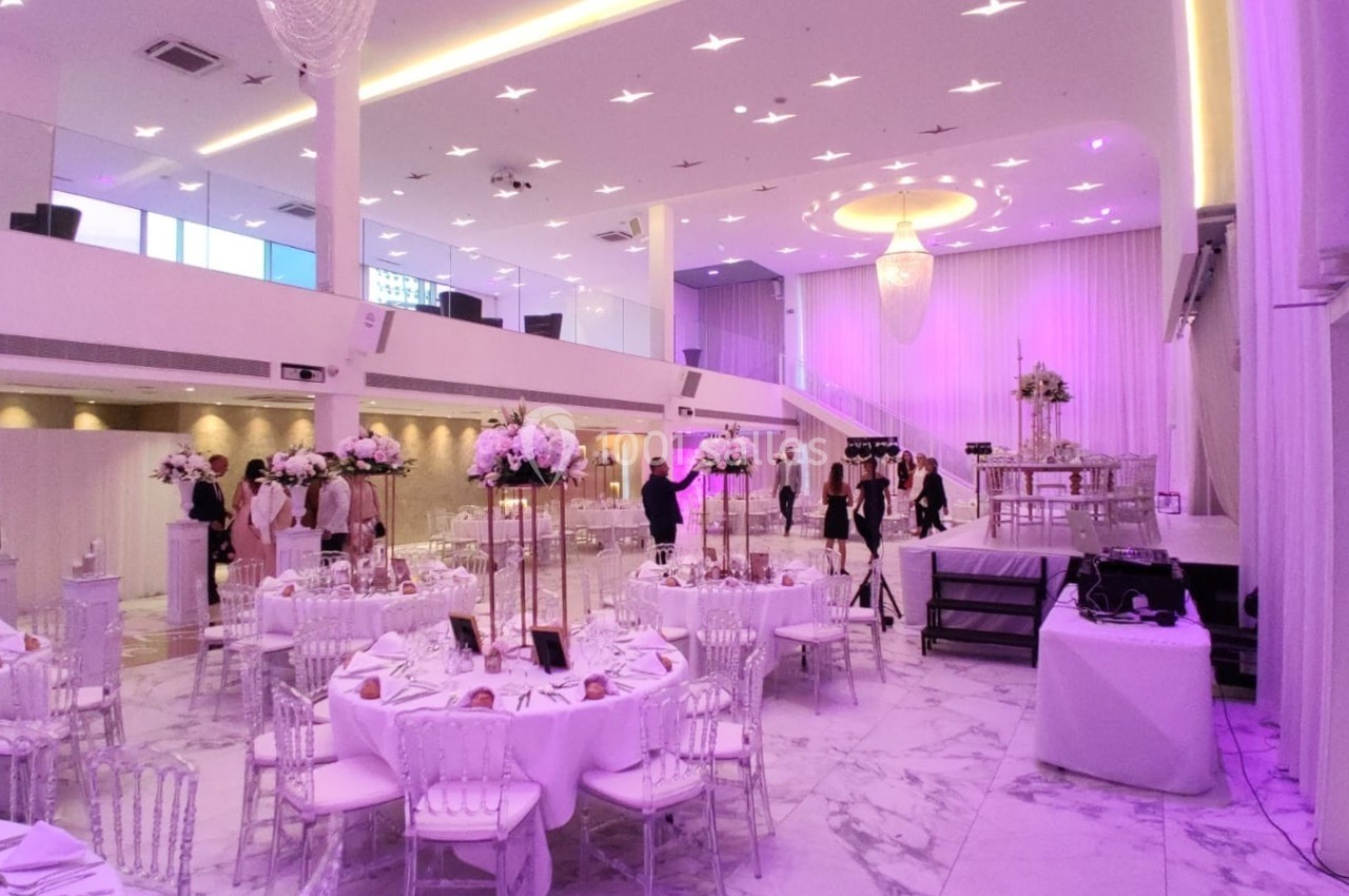 Salle de réception élégante avec tables décorées de fleurs, éclairage violet et espace scénique en arrière-plan.