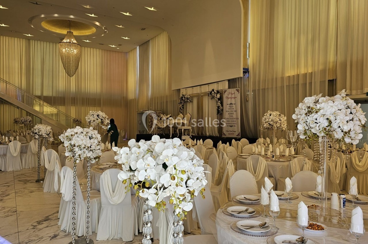 Salle de réception élégante décorée de fleurs blanches, avec tables dressées et grand lustre suspendu.