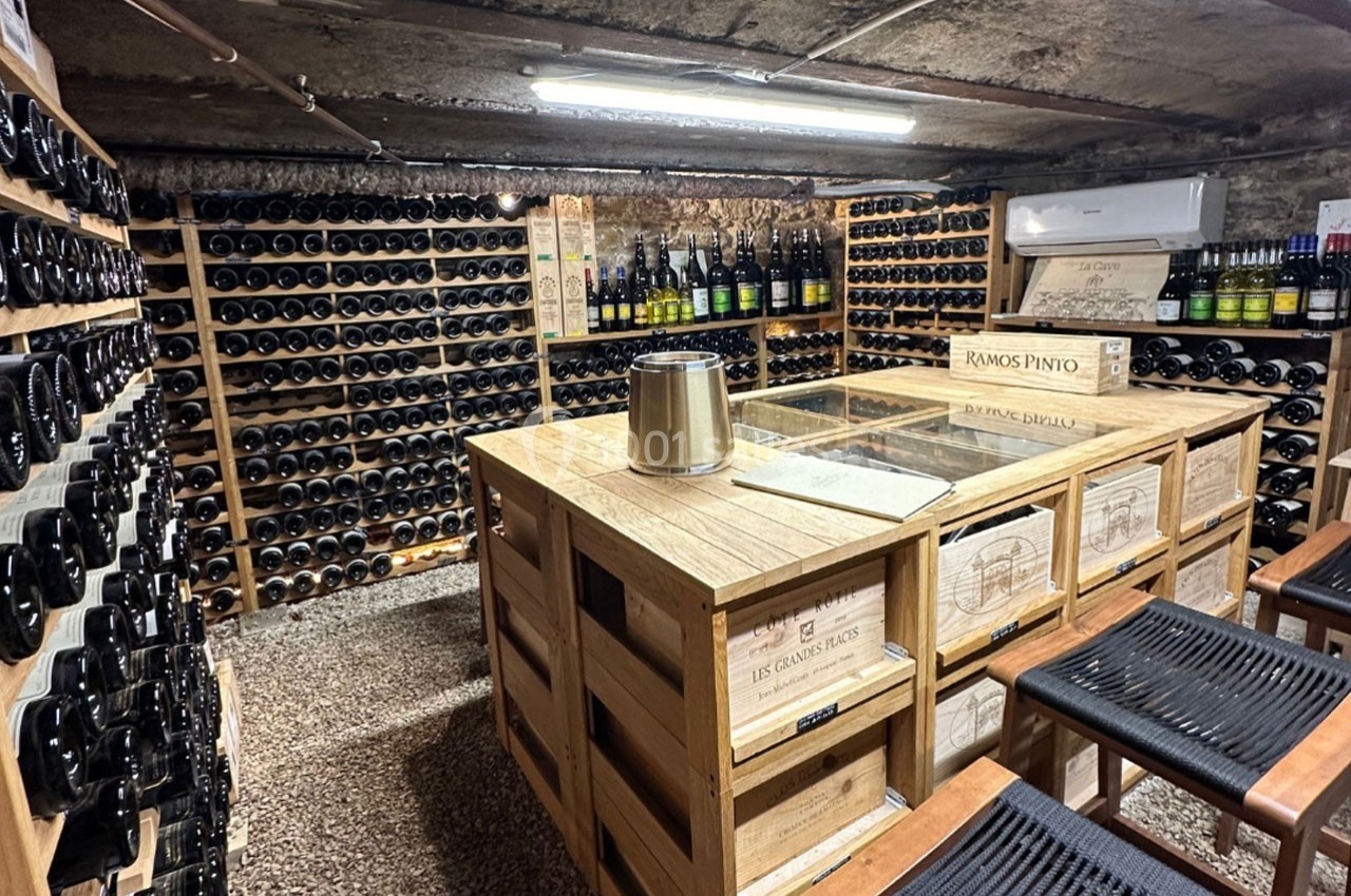 Cave à vin avec étagères remplies de bouteilles, table en bois au centre et tabourets en premier plan.