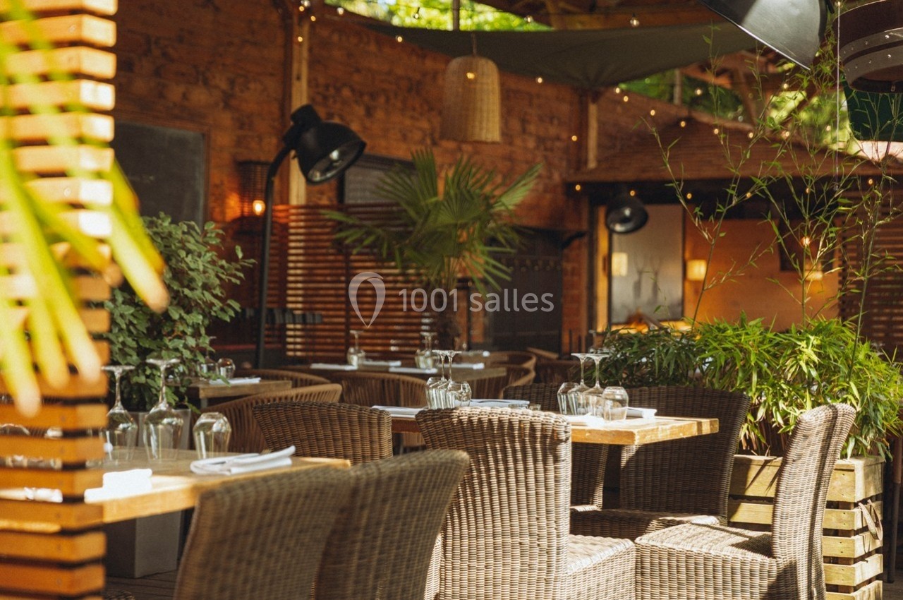 Terrasse de restaurant avec tables en bois, chaises en rotin, plantes et éclairage tamisé sous un auvent.