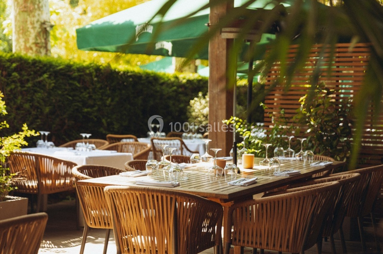Terrasse extérieure avec des tables en bois, chaises en rotin, couverts dressés et parasols verts sous un éclairage naturel.