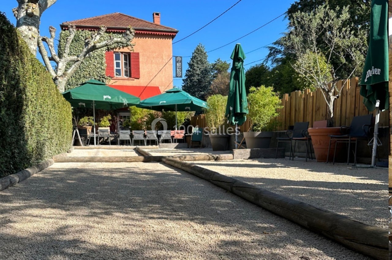 Terrasse extérieure avec terrain de pétanque, parasols verts, chaises, et maison rouge en arrière-plan.
