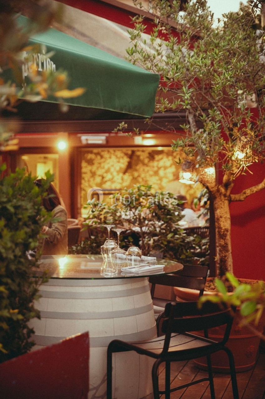 Table ronde en verre posée sur un tonneau, entourée de chaises noires, dans une terrasse extérieure arborée.
