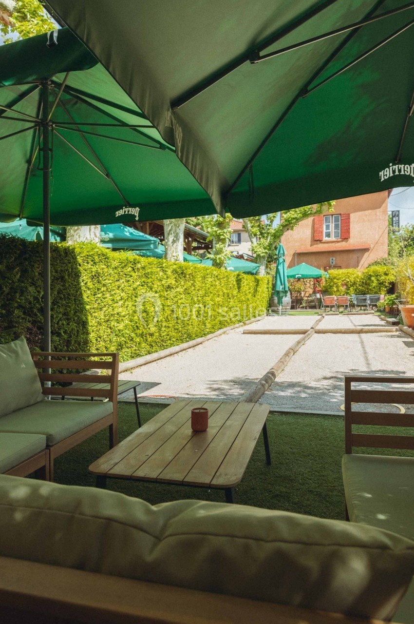 Espace extérieur d'un café avec tables, chaises et parasols verts, entouré de haies et d'arbres.