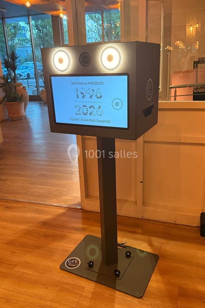 Borne photobooth sur pied avec écran tactile allumé, située dans un espace intérieur lumineux avec sol en bois.