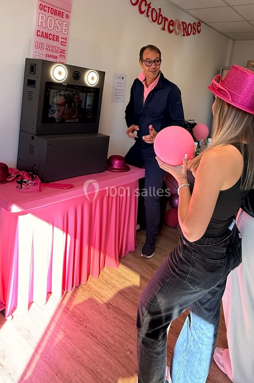 Deux personnes interagissent près d'une borne photo dans une salle décorée pour Octobre Rose, avec des accessoires roses.