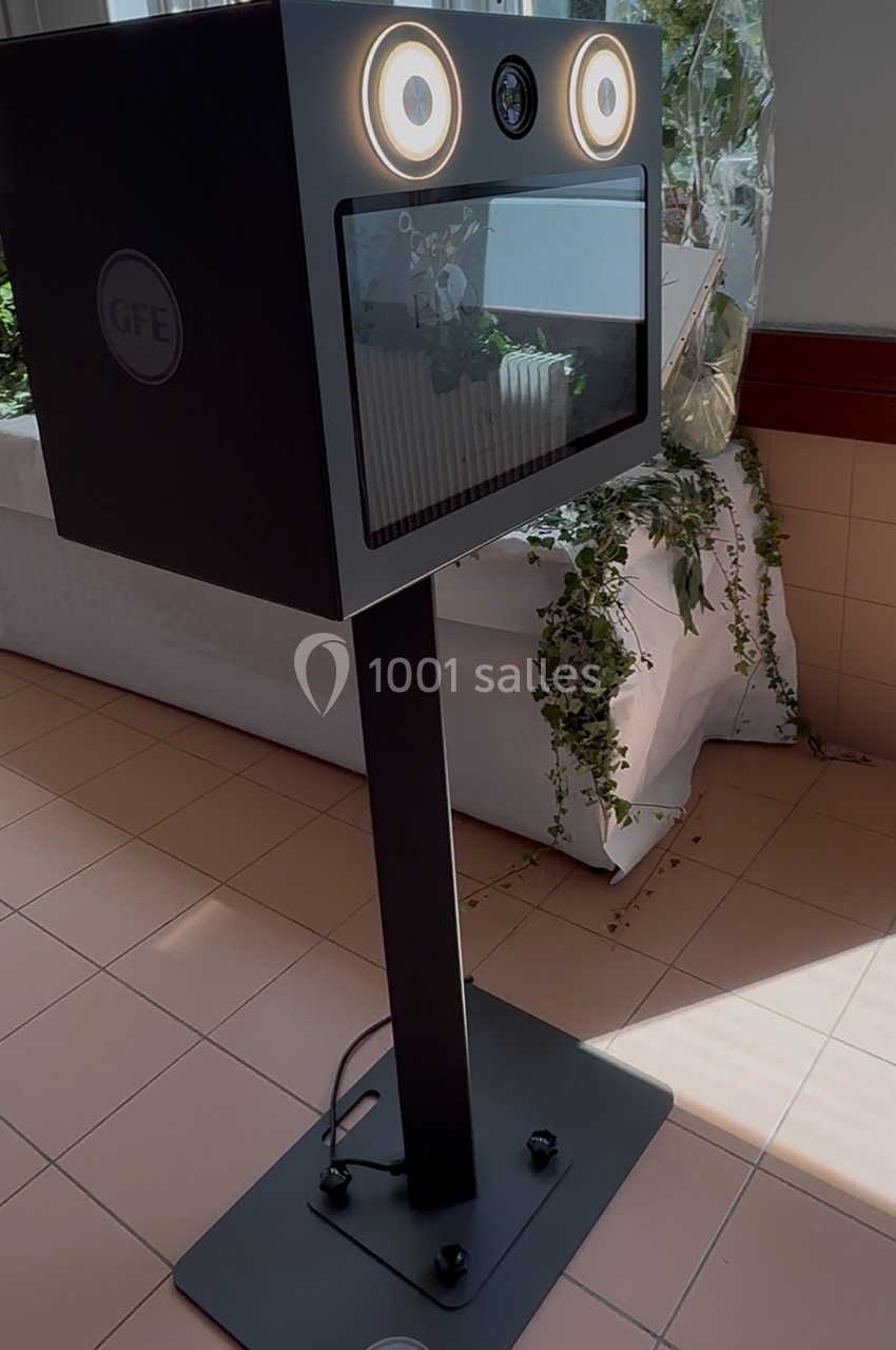 Borne photo noire sur pied avec éclairage circulaire, installée près d'une fenêtre et entourée de plantes décoratives.
