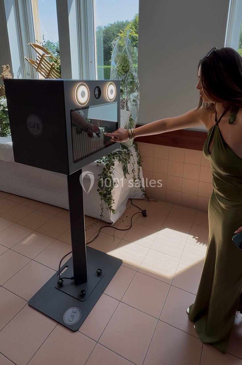 Une femme interagit avec une borne photo moderne dans un espace lumineux décoré de plantes.