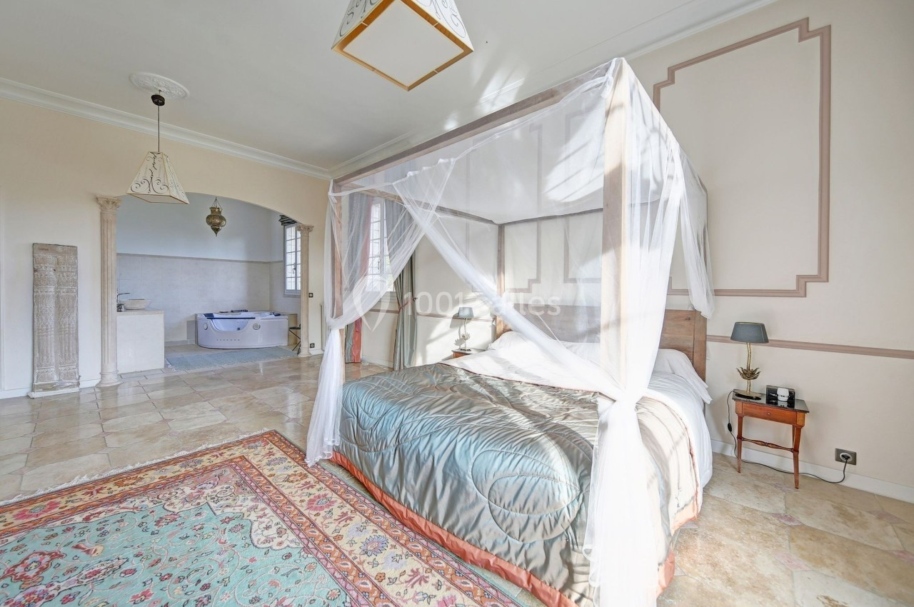 Chambre lumineuse avec lit à baldaquin, moustiquaire, tapis coloré et vue sur une salle de bain avec baignoire.