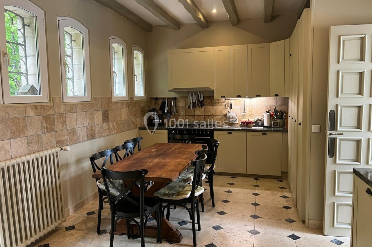 Cuisine lumineuse avec table en bois, chaises noires, meubles beige clair, carrelage clair et fenêtres en arc.