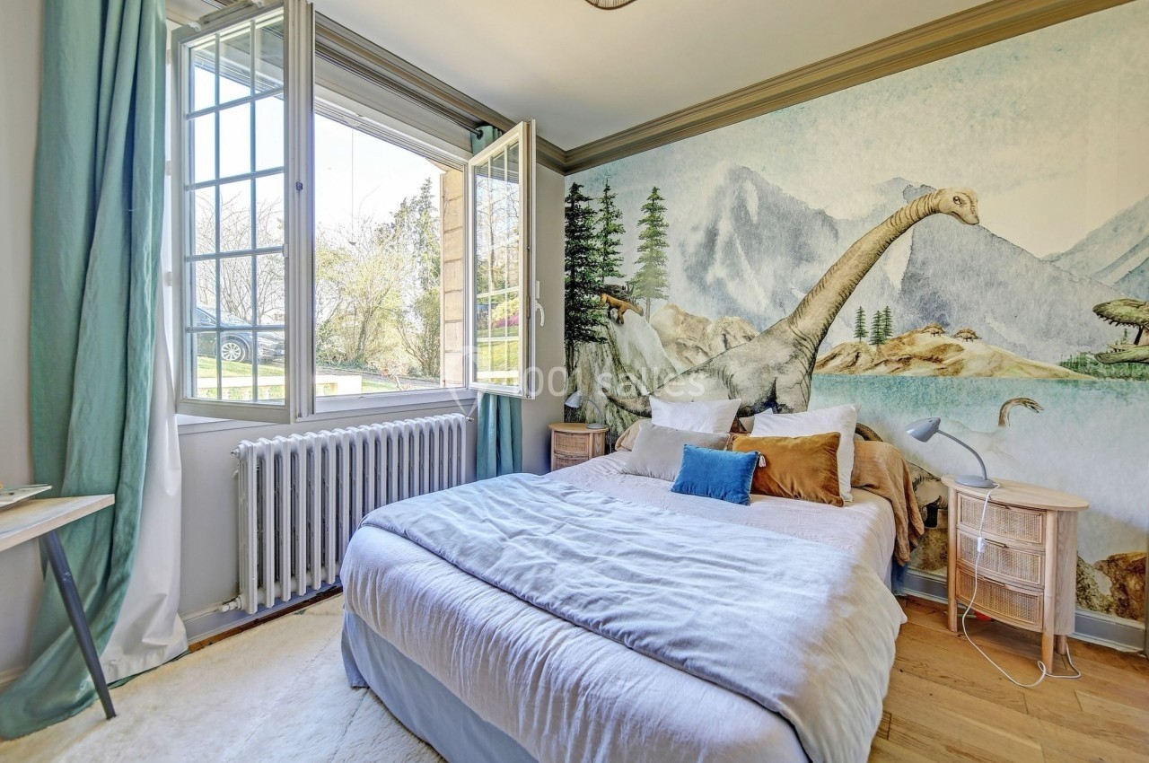 Chambre lumineuse avec lit double, grande fenêtre ouverte et mur décoré d'une fresque représentant des dinosaures.