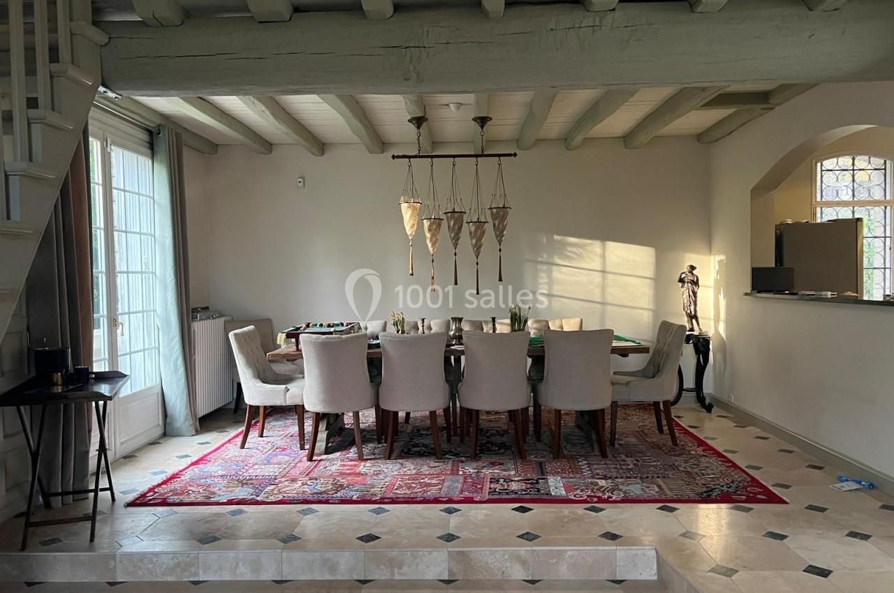 Salle à manger lumineuse avec une grande table entourée de chaises, tapis rouge au sol et poutres apparentes au plafond.