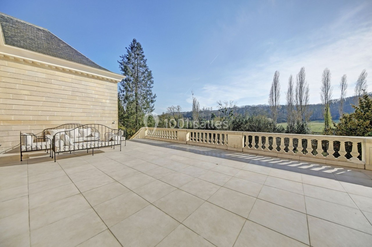Grande terrasse en pierre avec balustrade, mobilier en fer forgé et vue dégagée sur un paysage arboré.