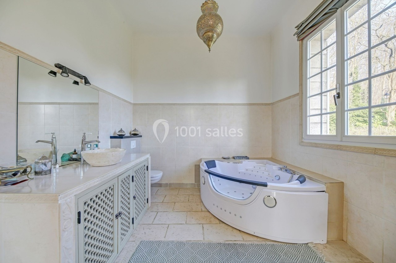 Salle de bain lumineuse avec baignoire d'angle à jets, vasque en pierre, grand miroir et fenêtre donnant sur un jardin.
