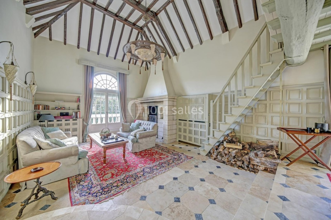 Salon lumineux avec poutres apparentes, cheminée en pierre, escalier en bois et mobilier classique sur tapis coloré.