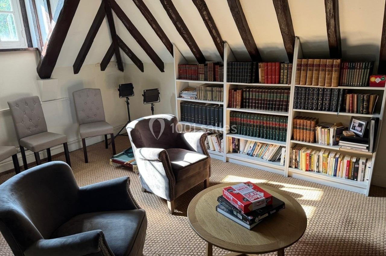 Salle de lecture avec fauteuils, table basse, bibliothèque garnie de livres et poutres apparentes au plafond.