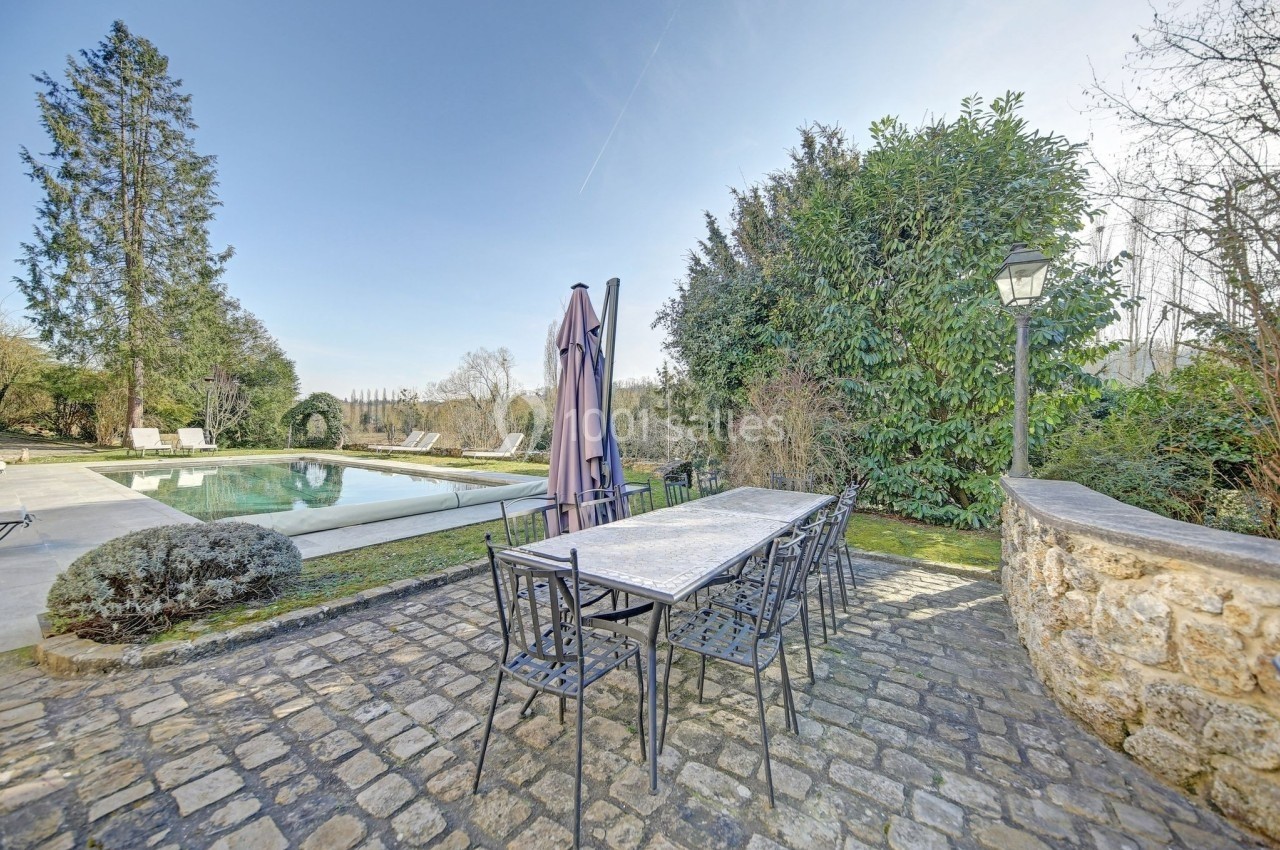 Table et chaises en métal sur une terrasse pavée, près d'une piscine entourée de végétation et d'arbres.