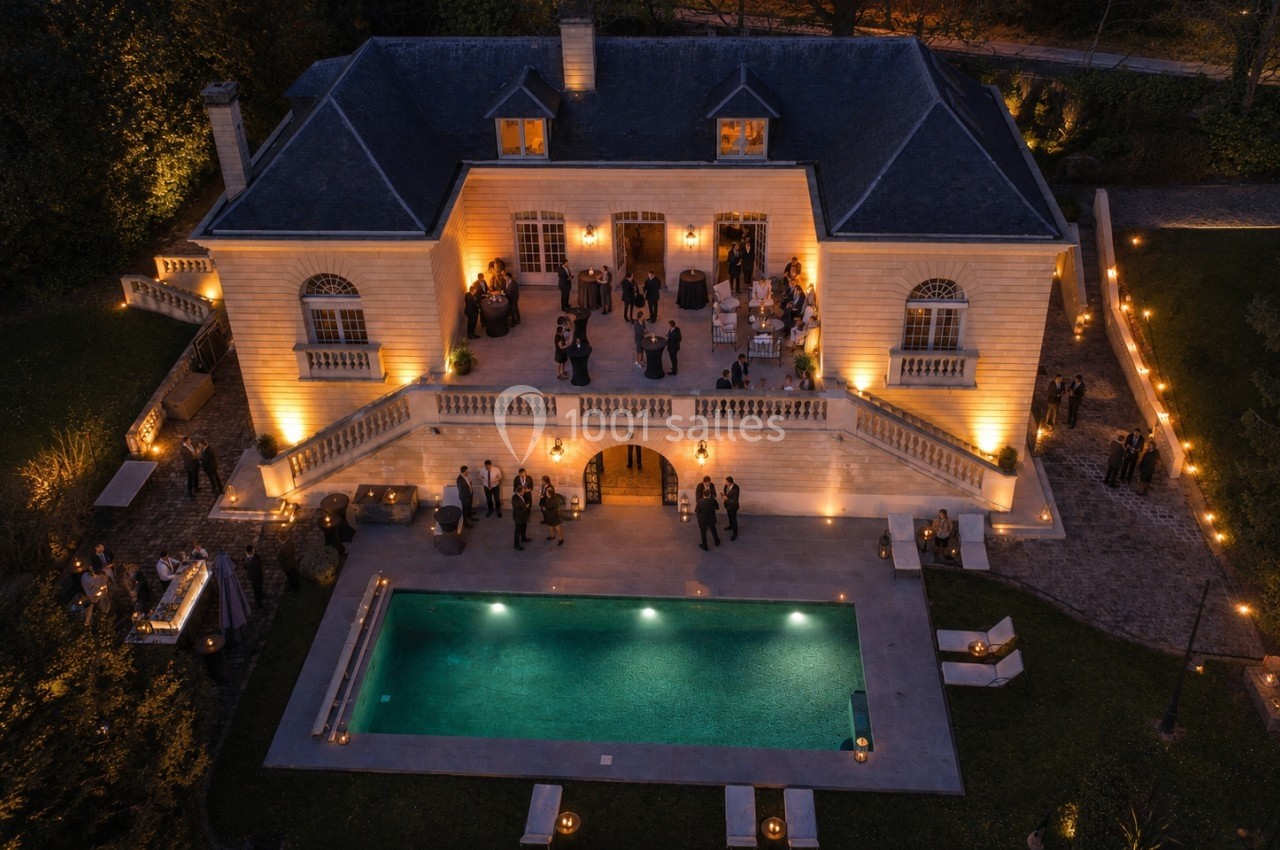 Vue aérienne d'une maison éclairée en soirée avec une piscine et des invités rassemblés sur la terrasse et dans le jardin.