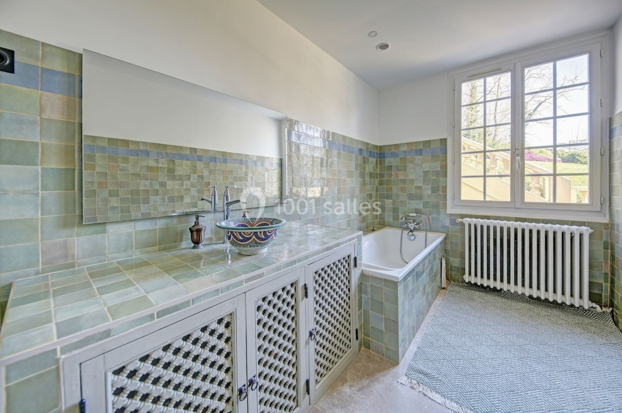 Salle de bain lumineuse avec carrelage vert, vasque décorative, baignoire, radiateur et fenêtre donnant sur l'extérieur.