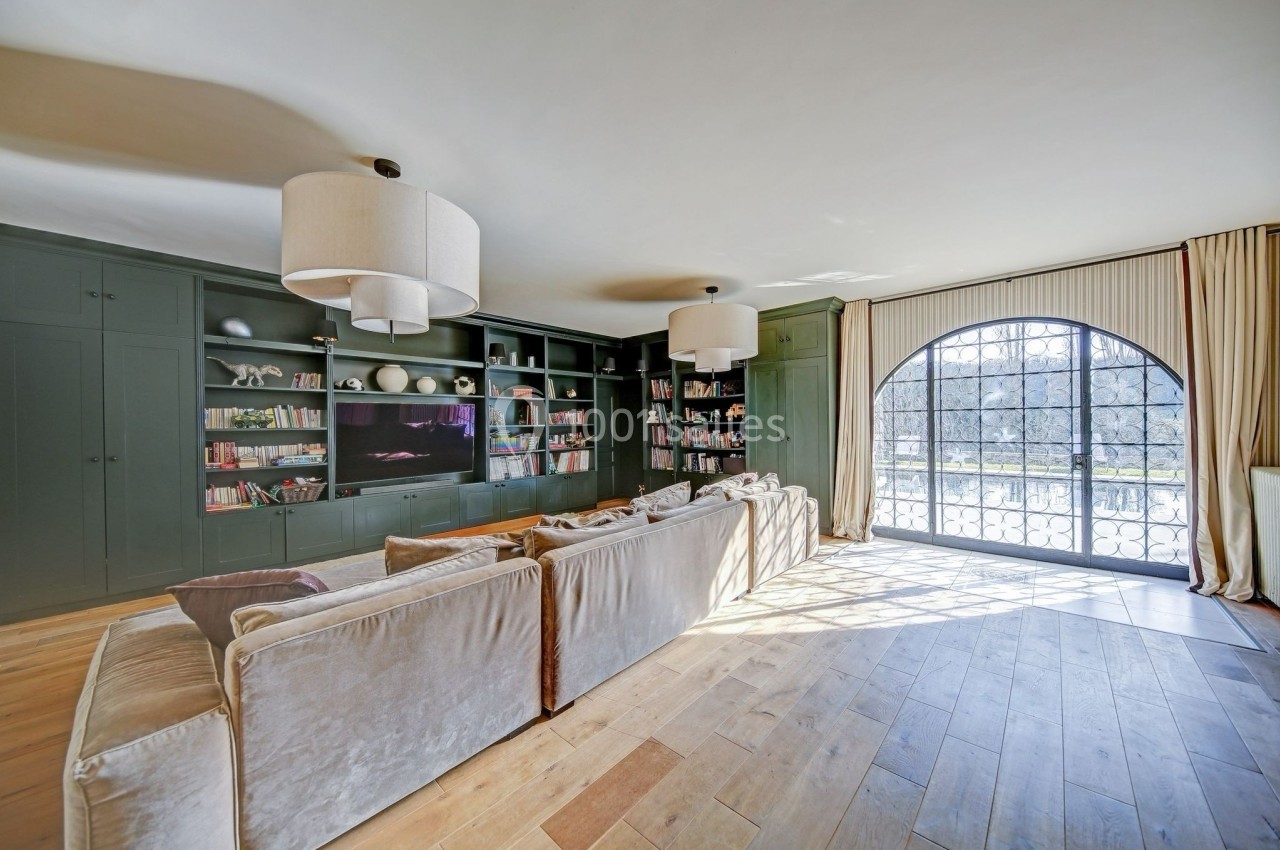 Salon lumineux avec canapé beige, bibliothèque murale verte, parquet en bois clair et grande baie vitrée arrondie.