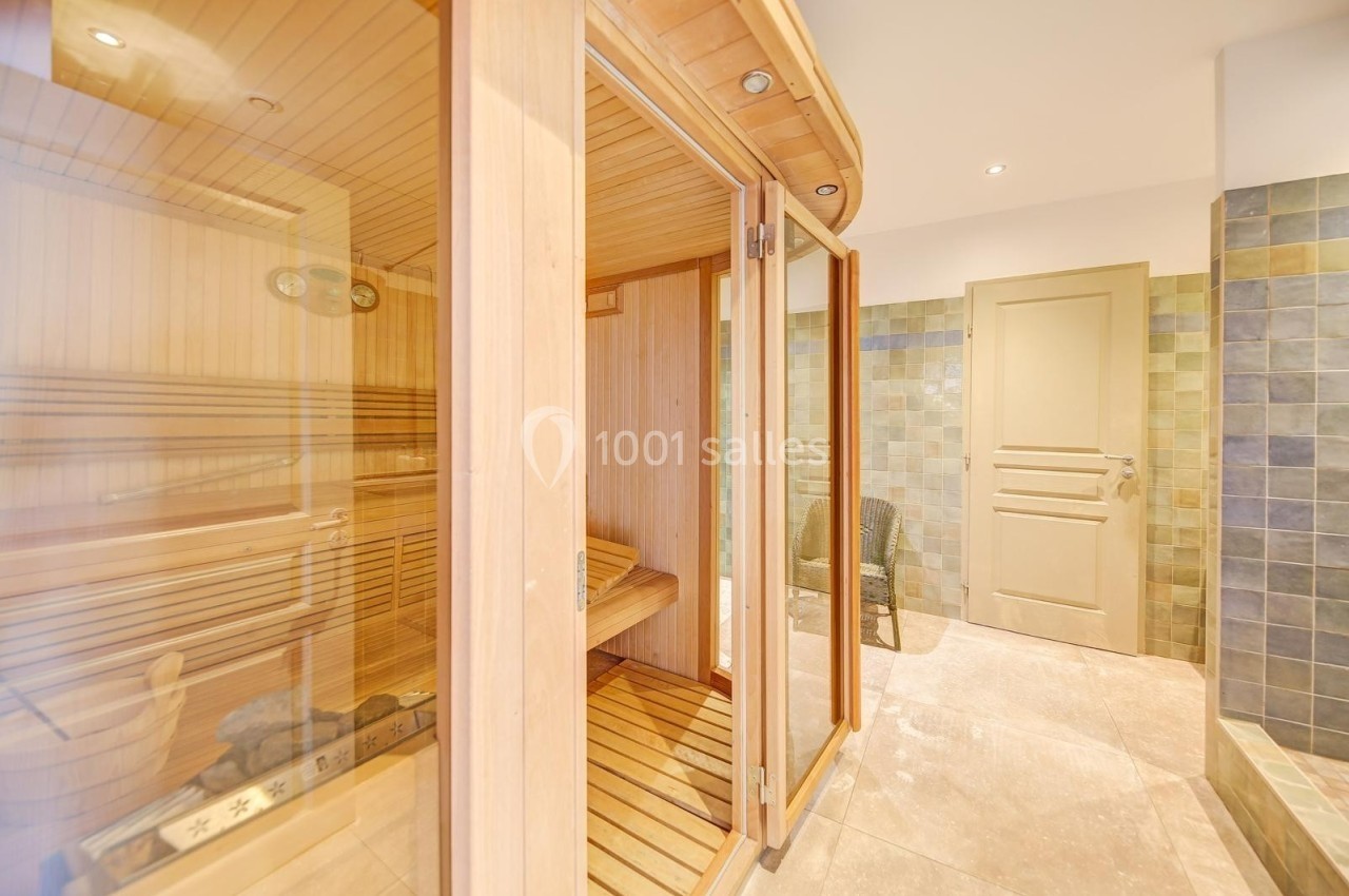 Sauna en bois avec porte vitrée, situé dans une pièce lumineuse aux murs carrelés et sol en pierre beige.
