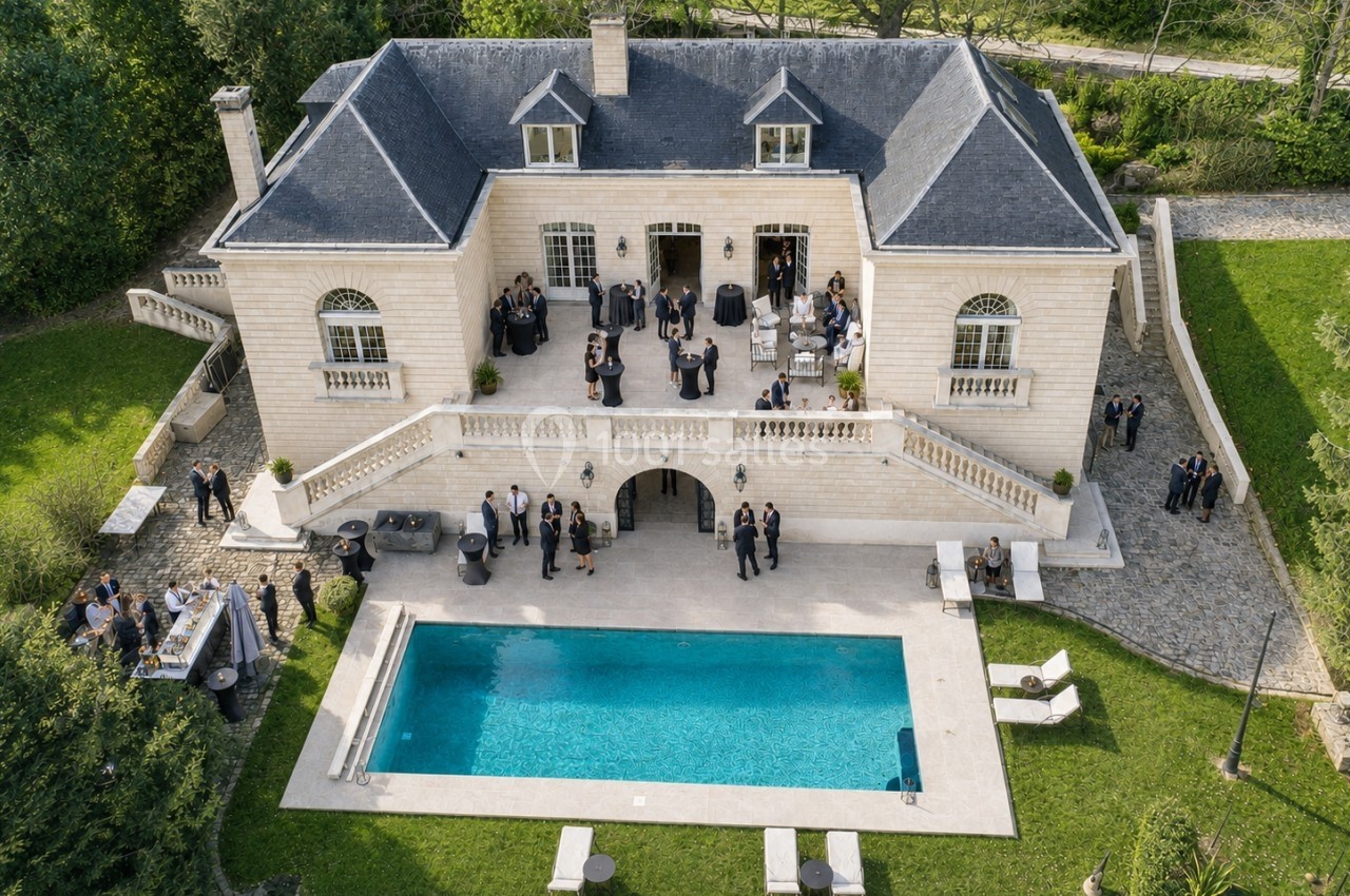 Vue aérienne d'une grande maison avec piscine, entourée de verdure, où des personnes sont rassemblées lors d'un événement.