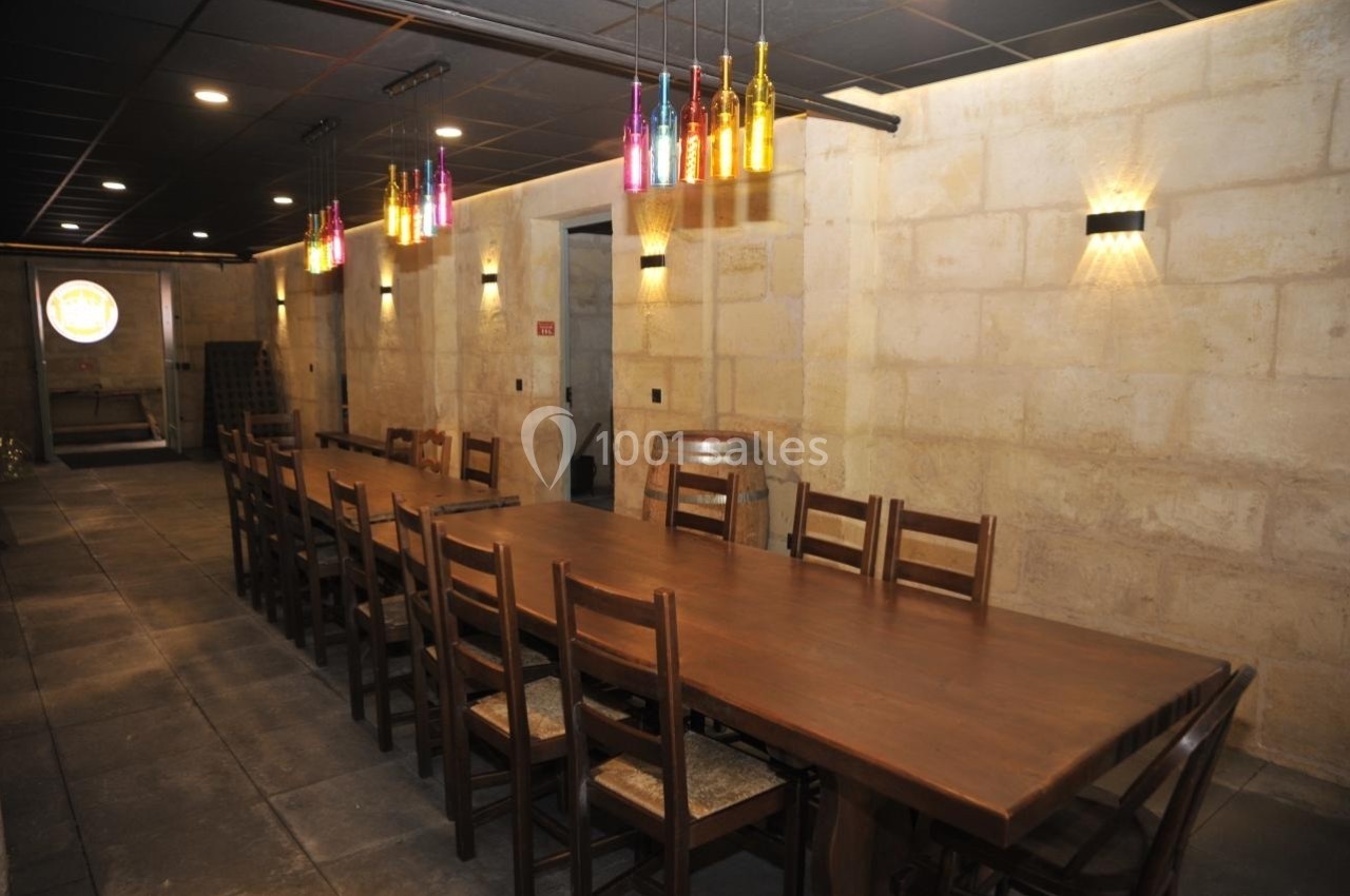 Salle en pierre avec une longue table en bois, des chaises assorties et des bouteilles colorées suspendues en éclairage.
