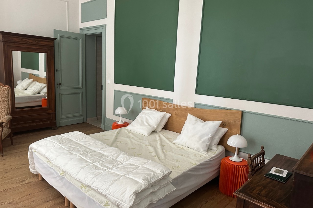Chambre avec lit double, murs verts, mobilier en bois, miroir sur pied et lampes de chevet orange.