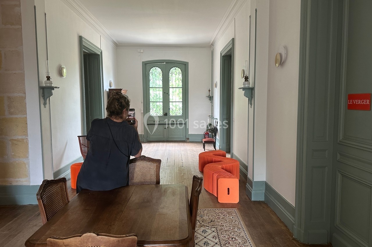 Une femme de dos prend une photo dans une pièce lumineuse avec des meubles en bois et des poufs orange.