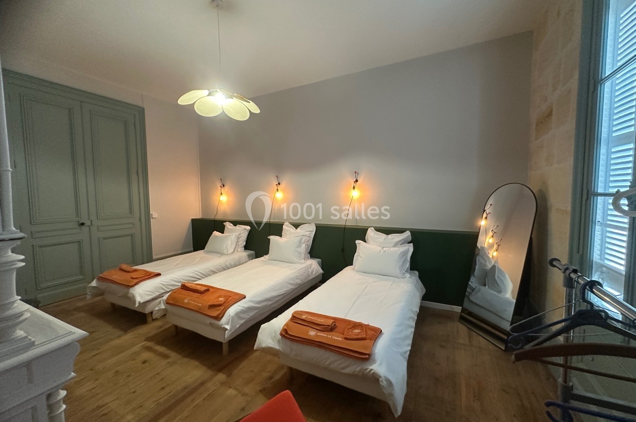 Trois lits simples avec linge de lit blanc et couvertures orange dans une chambre lumineuse au décor épuré.