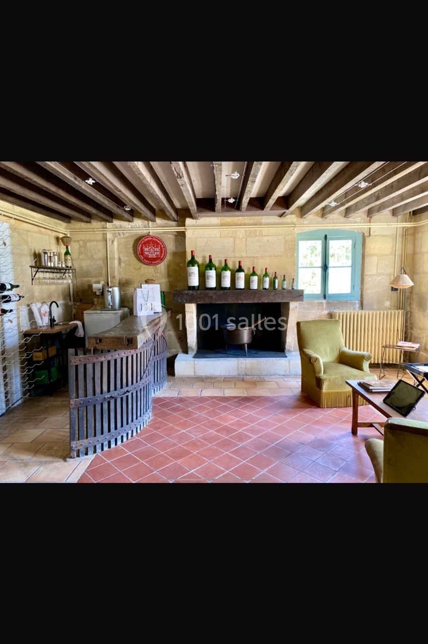 Salle rustique avec comptoir en bois, cheminée décorée de bouteilles, fauteuils et lumière naturelle.